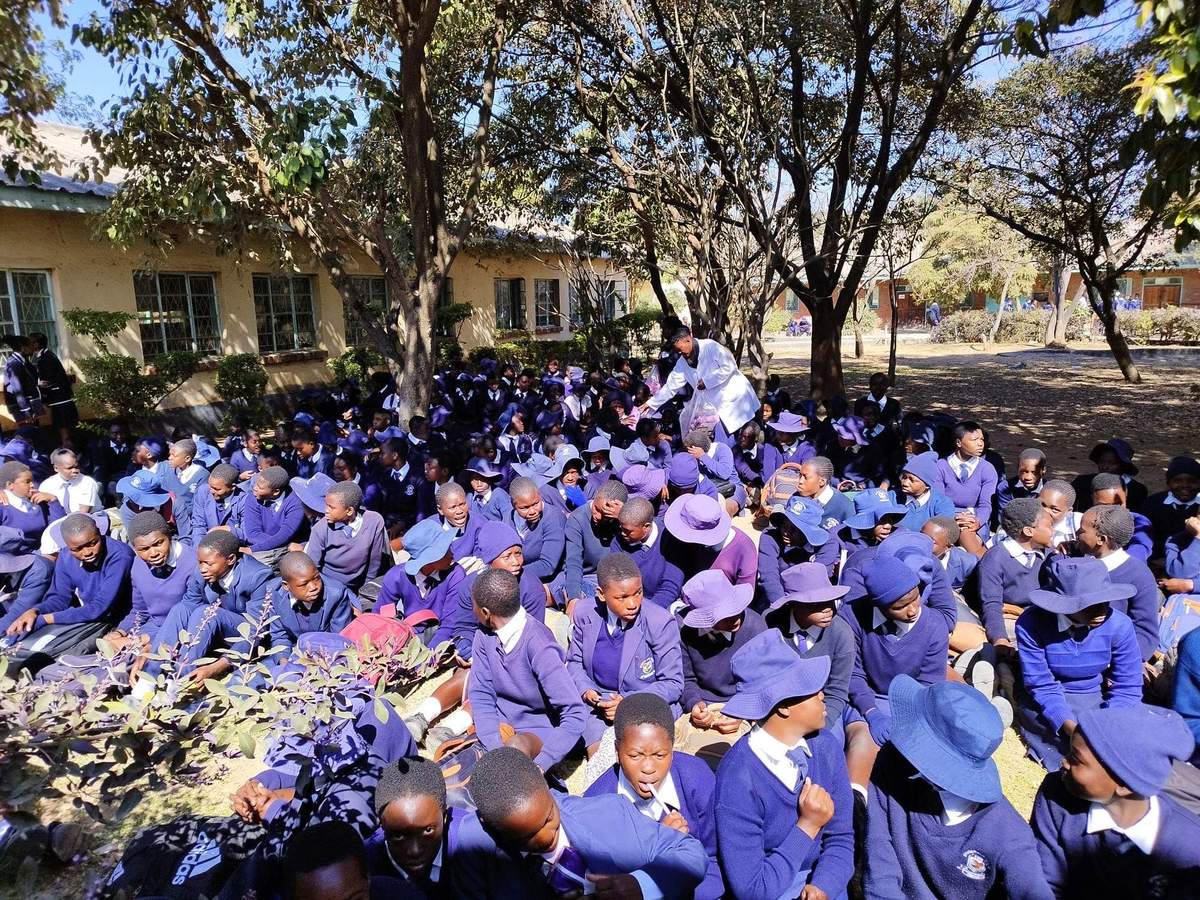 Large group of schoolchildren in purple and blue uniforms seated outdoors near a school building